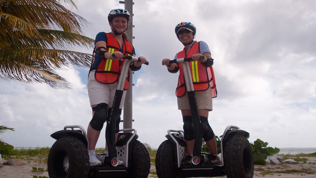 Dos mujeres con rodilleras, chalecos de seguridad y cascos sonríen mientras montan Segways Dos mujeres con rodilleras, chalecos de seguridad y cascos sonríen mientras montan Segways