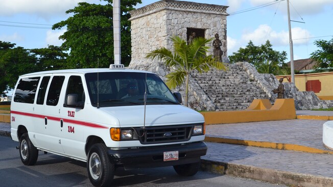 Uma van com uma placa de táxi no teto estacionada ao lado de uma palmeira e de um antigo edifício de pedra