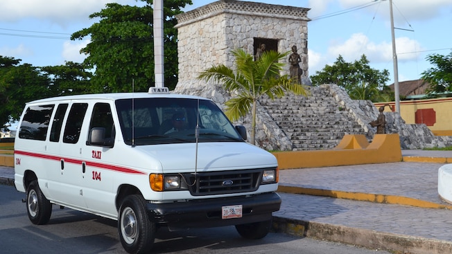 Furgoneta, con un cartel de taxi en el techo, estacionada junto a una palmera y un edificio de piedra de aspecto antiguo