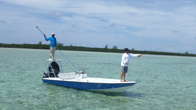 A man paddles a flats boat from a perch, while another man casts a line from a fishing pole A man paddles a flats boat from a perch, while another man casts a line from a fishing pole