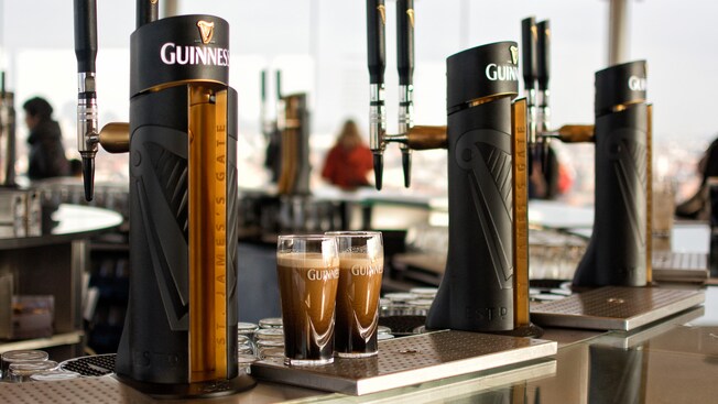 2 full Guinness logo glasses of dark beer amid kegs that also bear the Guinness logo