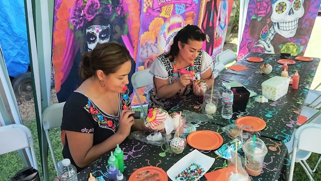 2 women decorate skulls at a table covered in crafting material 2 women decorate skulls at a table covered in crafting material