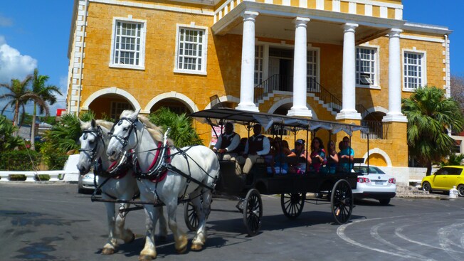 Two horses pulling a buggy filled with several passengers alongside a stone colonial structure