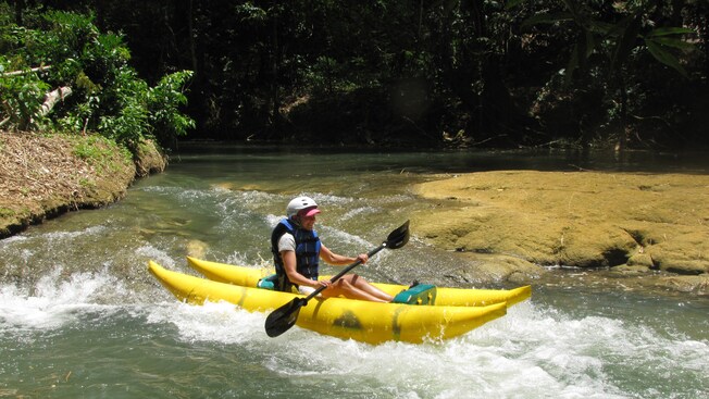 An adolescent outfitted in a life jacket and helmet navigates a single person kayak through mild white water waves