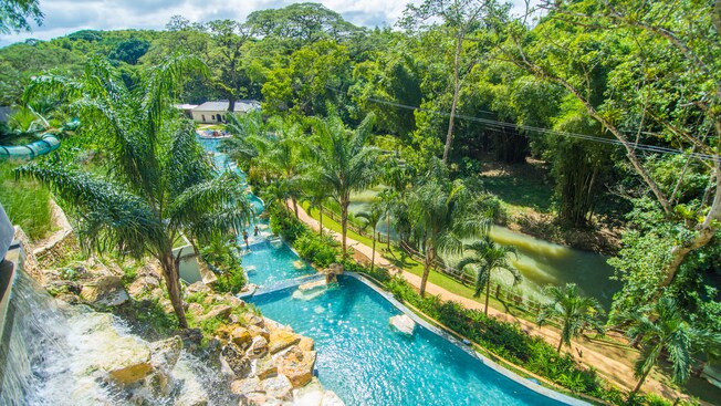 Una familia se encuentra en una piscina en cascada junto a árboles, una cascada, un tobogán, edificios, un río y rocas