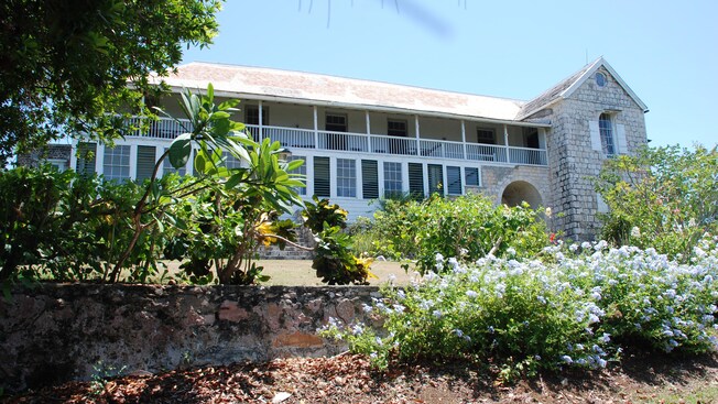 Tropical foliage fronts the entrance of 2 story stone plantation home with a verandah on second floor