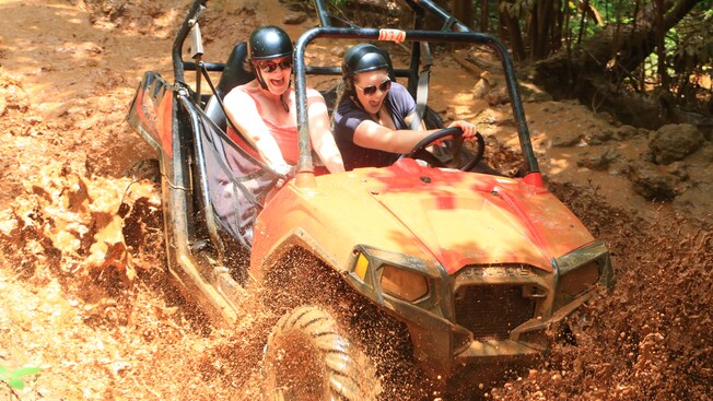 Mulheres usando óculos escuros e capacetes sorriem enquanto dirigem por um terreno muito lamacento em um buggy