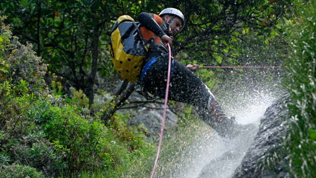 Man with a helmet and backpack holds on to a rope as he repels down a waterfall in the forest