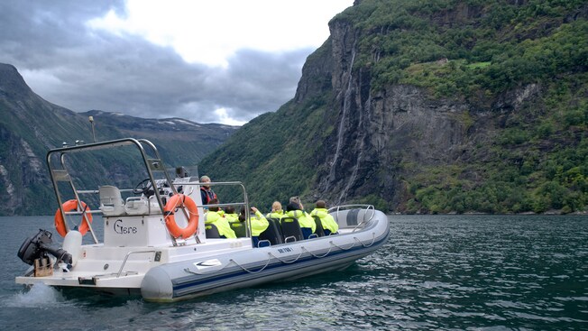 People sitting in an inflatable boat facing a steep canyon wall with vegetation and waterfalls