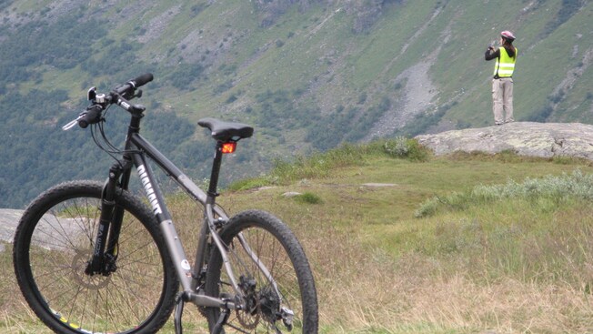A woman stands on a rock taking a picture of a grassy hillside with a bicycle in the foreground
