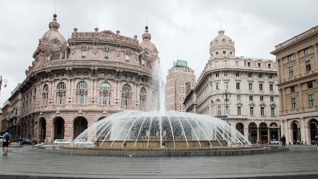 People walk near a fountain and old city buildings People walk near a fountain and old city buildings