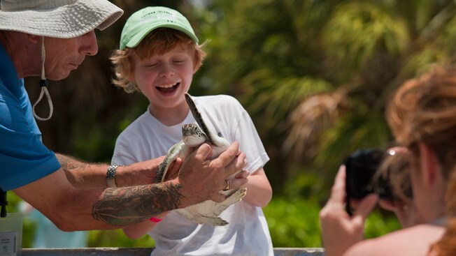 A guide hands a small smiling boy a young sea turtle while a woman nearby photographs the moment