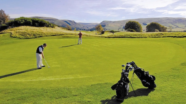 Male golfer #1 lines up his golf ball while male golfer #2 holds the pin