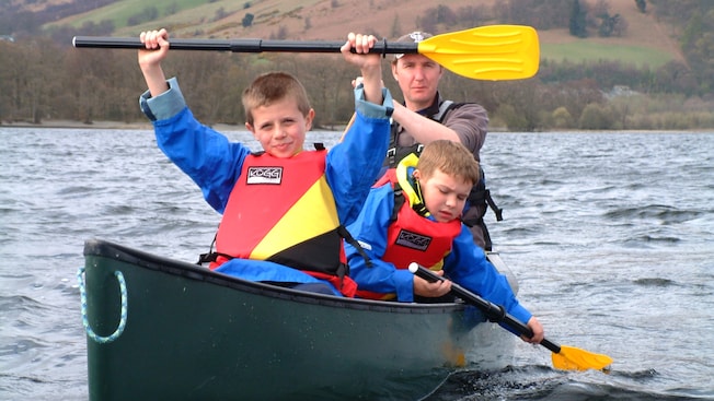 Older brother holds up an oar over his head while his younger brother and father paddle the canoe