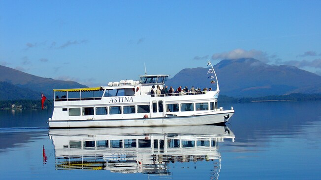 A boat with 2 decks carrying passengers across a large body of water with hills in the distance