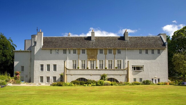 A large building featuring windows, chimneys, trees, flowers and a lawn