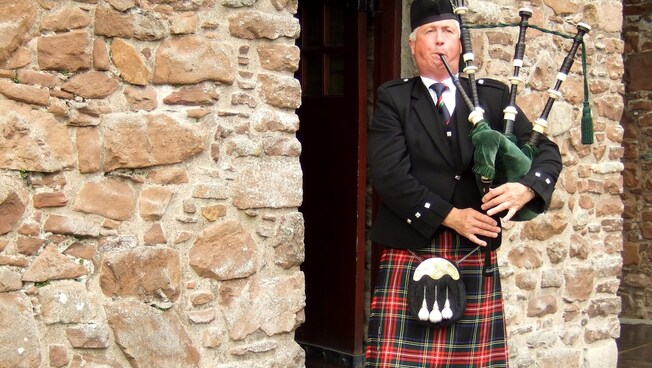 A man wearing a kilt plays bagpipes in the doorway of an old stone building
