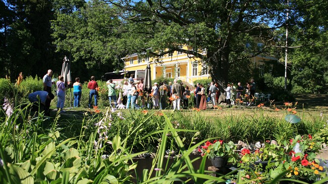 A tour group listens to a guide outside an old manor surrounded by old growth trees and gardens