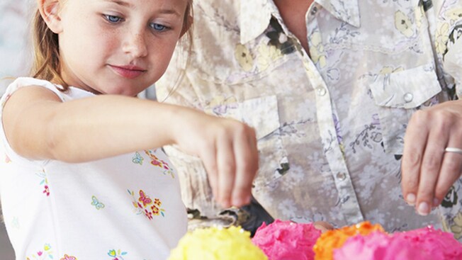 A little girl sprinkles toppings on a cupcake while her mom looks on