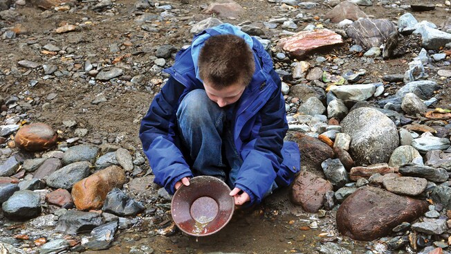 A young boy pans for gold on the rocky bank of a creek