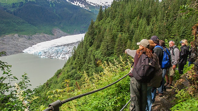 A group of people stand on a forest trail overlooking a valley and a glacier A group of people stand on a forest trail overlooking a valley and a glacier