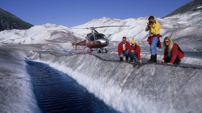 Four smiling people in light sweaters and jeans crouch along an ice ridge above a pool of glacier water