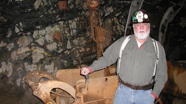 Bearded man with suspenders and miners hat stands in front of rusted mining equipment in a cave