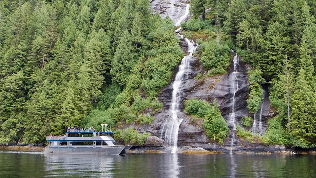 A ferry boat sails in front of a waterfall running off a mountain into the ocean