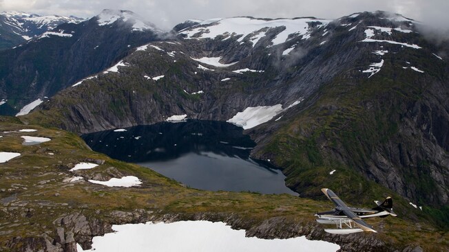 Un hidroavión volando sobre un paisaje montañoso con un lago glacial cerca de su cima