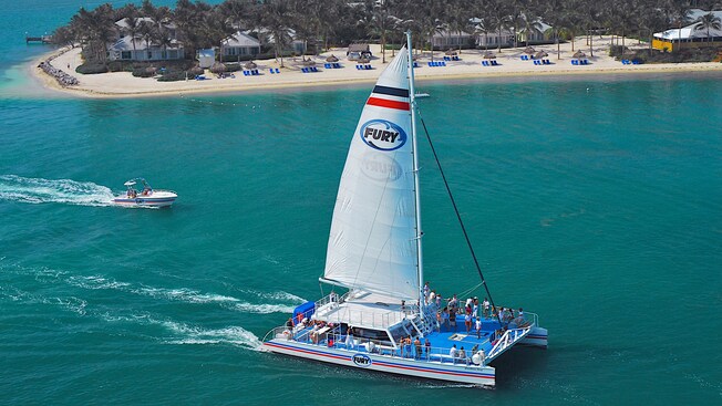 A group of Guests relaxing on the deck of a catamaran next to a shoreline containing palm trees, lounge chairs, palapa umbrellas and beach homes