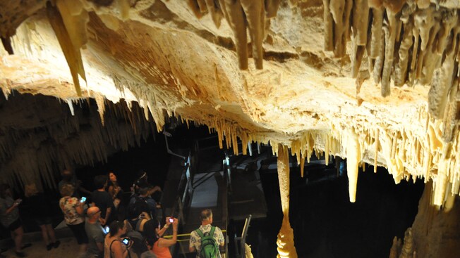 Stalactites hanging from the roof of a cave with visitors standing beneath Stalactites hanging from the roof of a cave with visitors standing beneath