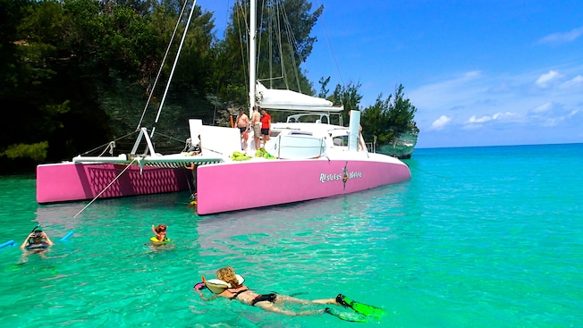 Um catamarã atracado em uma enseada, enquanto as pessoas mergulham de snorkel na água enquanto outras ficam de pé no convés Um catamarã atracado em uma enseada, enquanto as pessoas mergulham de snorkel na água enquanto outras ficam de pé no convés