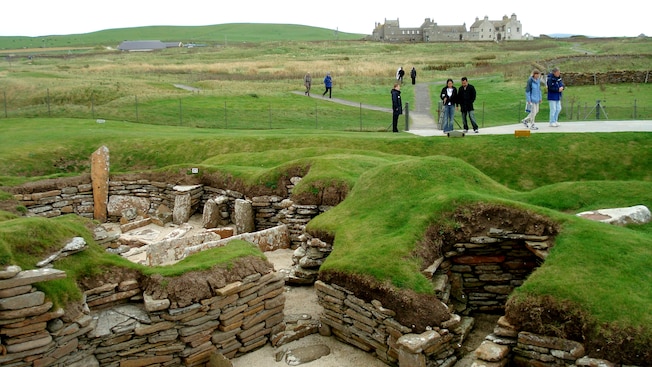 A Skaill House fica ao fundo enquanto os visitantes observam o assentamento da idade da pedra de Skara Brae A Skaill House fica ao fundo enquanto os visitantes observam o assentamento da idade da pedra de Skara Brae