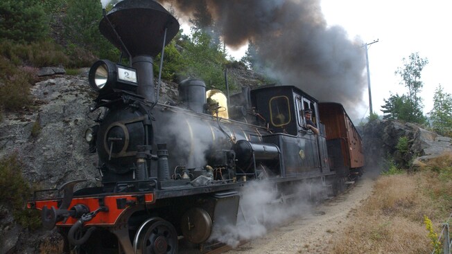 El conductor de un tren sonríe mientras saca la cabeza por la ventana de una maquina a vapor que se desplaza por terrenos montañosos