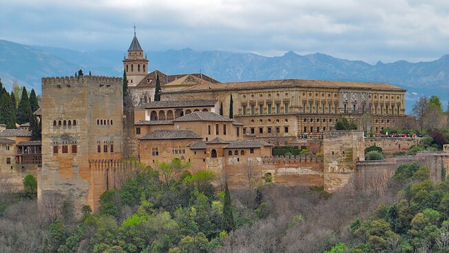 People walk around ramparts and a fortress near trees and distant mountains