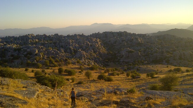 A woman stands in a clearing near rocks, bushes and distant mountains