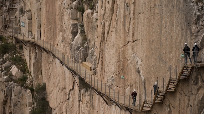People walk down a wooden path and stairs set into the side of a cliff People walk down a wooden path and stairs set into the side of a cliff