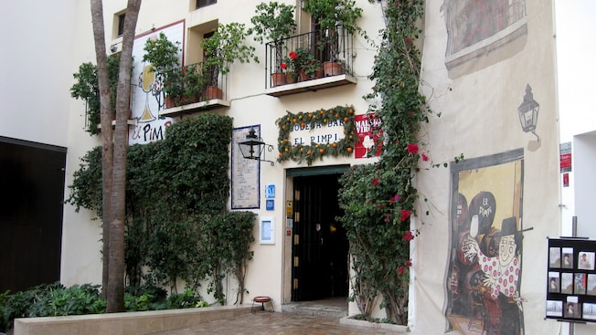 The front of a building near trees, bushes, vines and a sign that reads Bodega Bar El Pimpi The front of a building near trees, bushes, vines and a sign that reads Bodega Bar El Pimpi