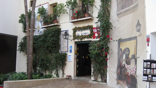 A frente de um edifício perto de árvores, arbustos, vinhas e uma placa que diz Bodega Bar El Pimpi
