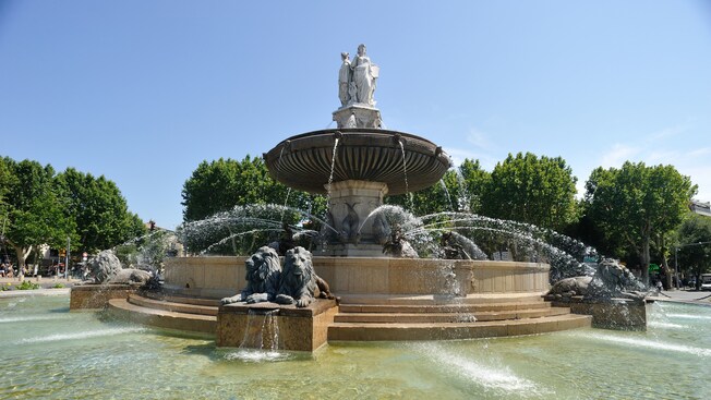 A fountain with statues and jets of water near trees and onlookers