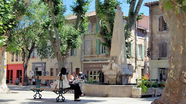 People sit by a fountain near trees, buildings and storefronts People sit by a fountain near trees, buildings and storefronts