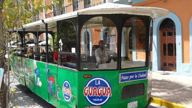 A trolley on a street featuring signs that say LA GUAGUA MAZATLAN, Paseo por la Ciudad