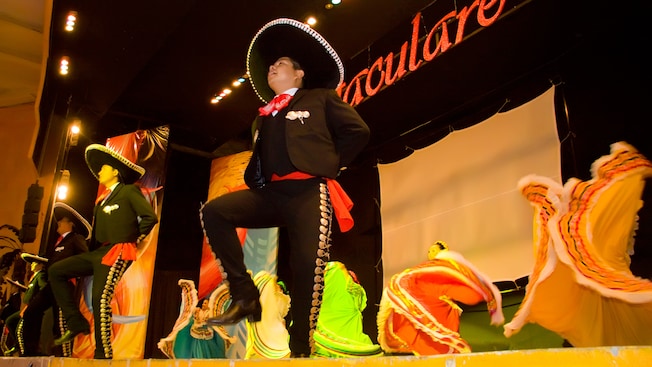 Performers in traditional Mexican attire dance on a stage with a sign that reads Spectaculare hanging above them Performers in traditional Mexican attire dance on a stage with a sign that reads Spectaculare hanging above them