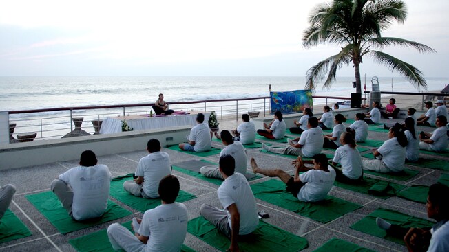 Facing a yoga instructor, Guests sit on yoga mats by the ocean and make the lotus yoga pose