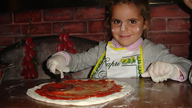 A small girl with flour on her face smiles as she spreads tomato paste over pizza dough A small girl with flour on her face smiles as she spreads tomato paste over pizza dough