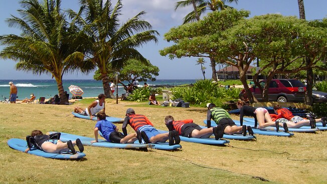 On a grass lawn in front of a beach, novice surfers practice pushing up from the paddling position