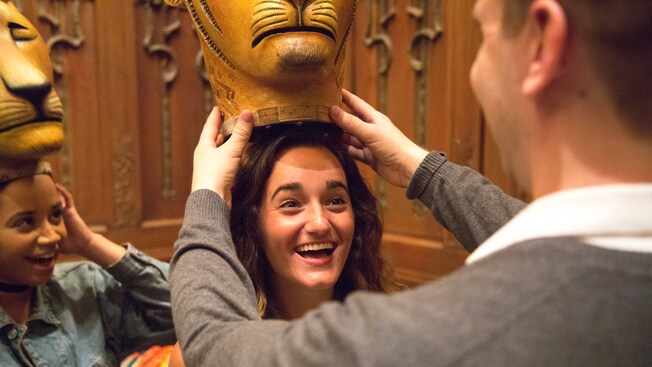 A woman smiles as a man puts a lion headdress on her head while another woman watches