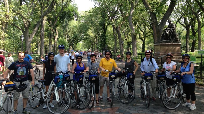 A group of Guests with bicycles smile into the camera on a paved, highly populated walkway in a large park