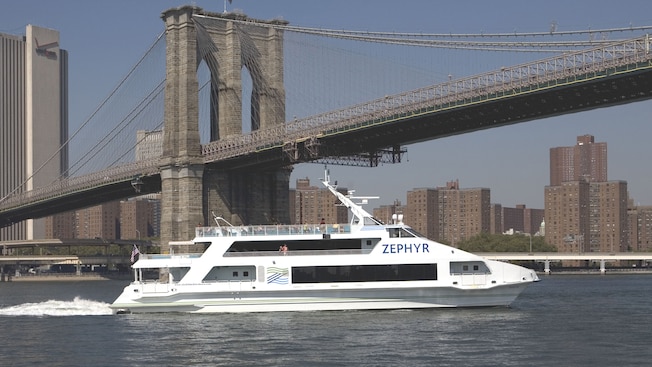 A yacht with travels along a river underneath a stone suspension bridge with high rises in the distance A yacht with travels along a river underneath a stone suspension bridge with high rises in the distance