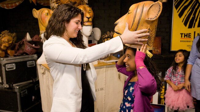 A woman helps a girl try on a lion mask in a room filled with The Lion King stage props